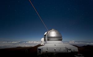 Dome of the Northern Gemini telescope high above the clouds. A beam of light is going up into a deep blue starry sky.