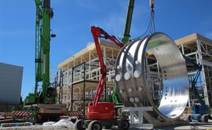 Three cranes lifting a large silver ring-shaped section of the chamber.