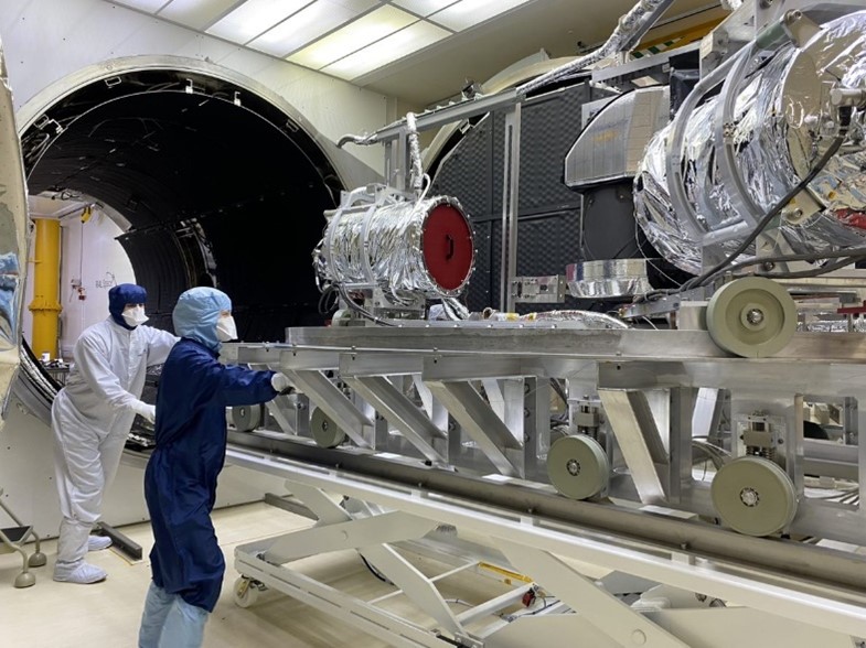 Two technicians in clean room suits roll the MWS PFM out of the chamber.