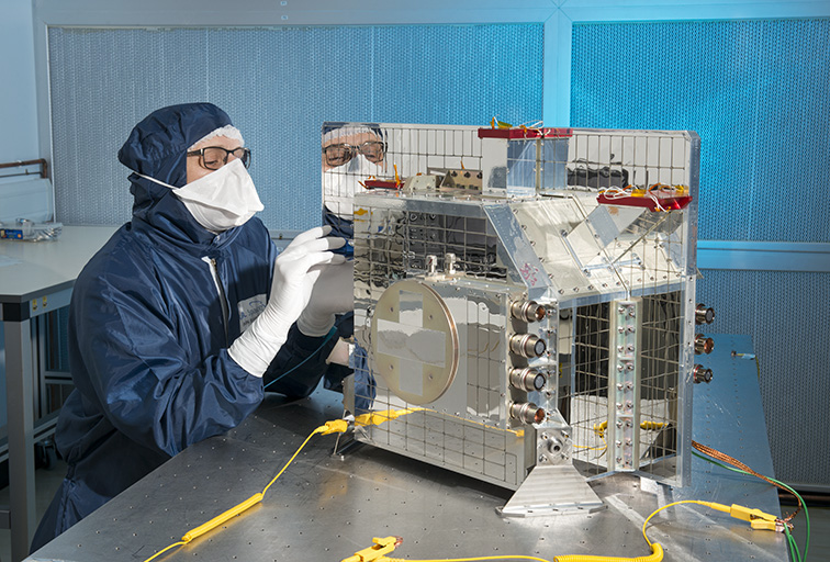 EarthCARE's BBR instrument in the lab at RAL Space. The instrument appears to be covered in mirrors, and an engineer wearing a clean room suit is focussing on a small part of it.
