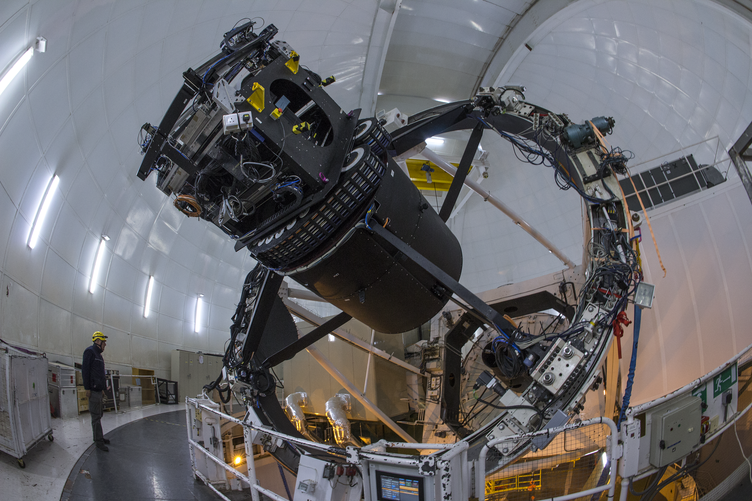 Inside the dome of the William Herschel Telescope, the WEAVE upgrade can be seen fixed to the telescope. A person can be seen standing in the frame, showing the impressive scale of the telescope itself.