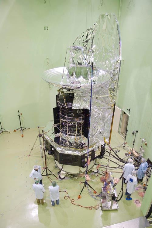 The Herschel Space Observatory in testing in the clean room with 6 people in clean suits inspecting.