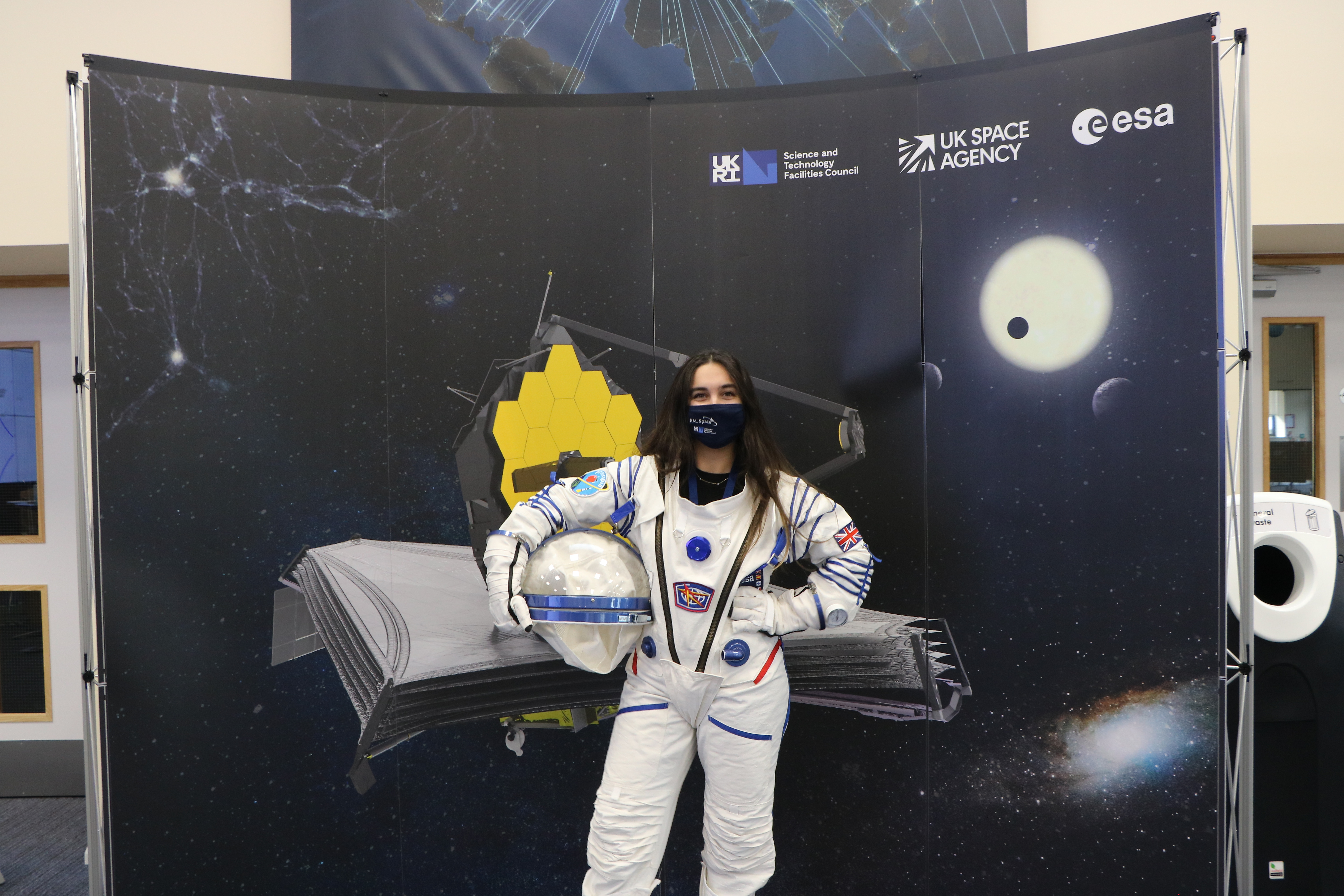 A young woman with long dark hair stands smiling wearing an astronaut suit and face mask.