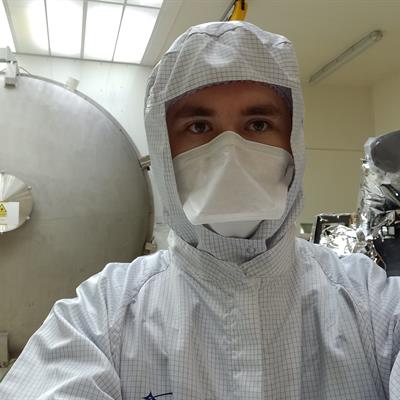 Ben Keeble wearing a clean room suit, standing outside of RAL Space's 3-metre space test chamber next to the SLSTR instrument.